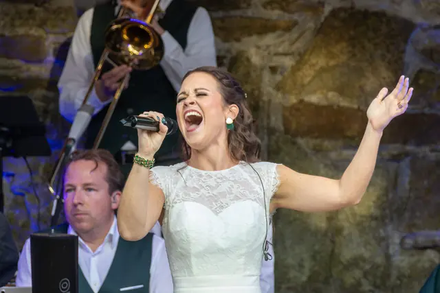 Sängerin im weißen Kleid singt mit ausgestrecktem Arm, hinter ihr Keyboard und Posaune.