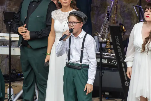 Junger Sänger mit Schiebermütze singt, neben ihm Sängerinnen in Weiß und Sänger in grünen Westen.