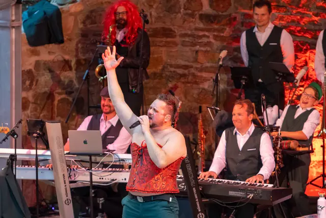Sänger im Korsett reckt den Arm in die Höhe, dahinter die Band an Keyboards und Bläsern.