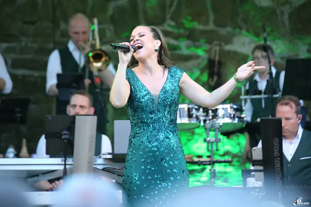 Eine Sängerin im grünen Paillettenkleid singt mit ausgebreitetem Arm, hinter ihr Posaune und Band.