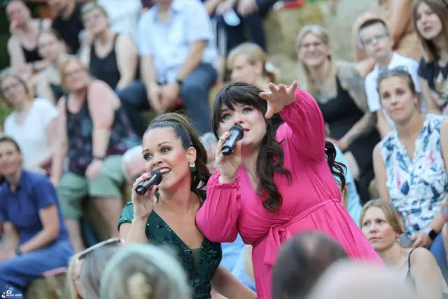 Zwei Sängerinnen, eine in Grün, eine in Pink, singen zwischen den Zuschauern auf den Rängen.