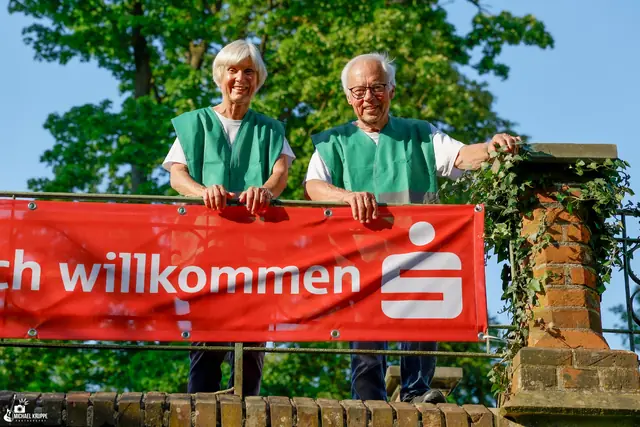 Zwei Helfer in grünen Westen stehen hinter einem Willkommens-Banner der Sparkasse.